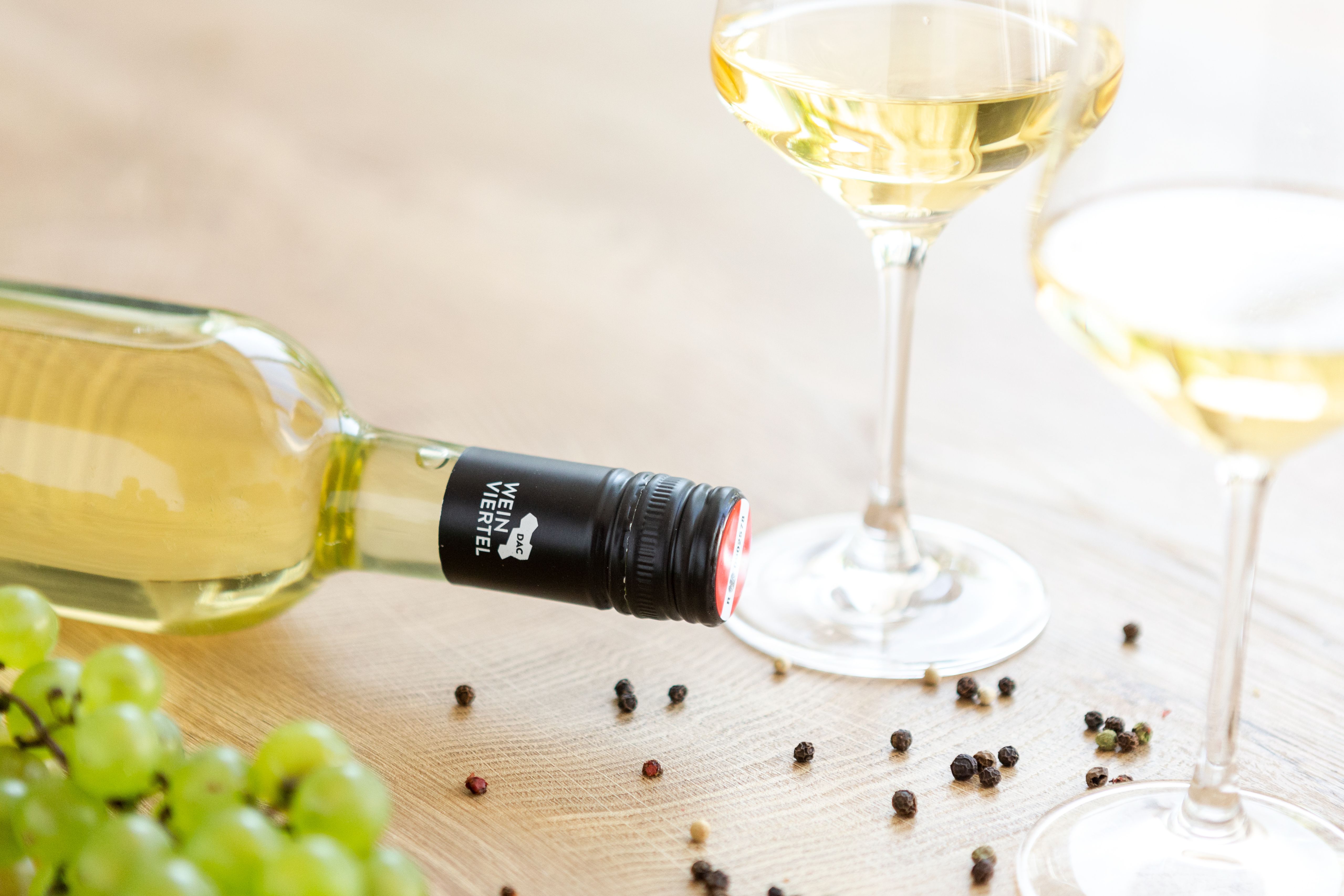 White wine bottle and glasses on a table with grapes and peppercorns.