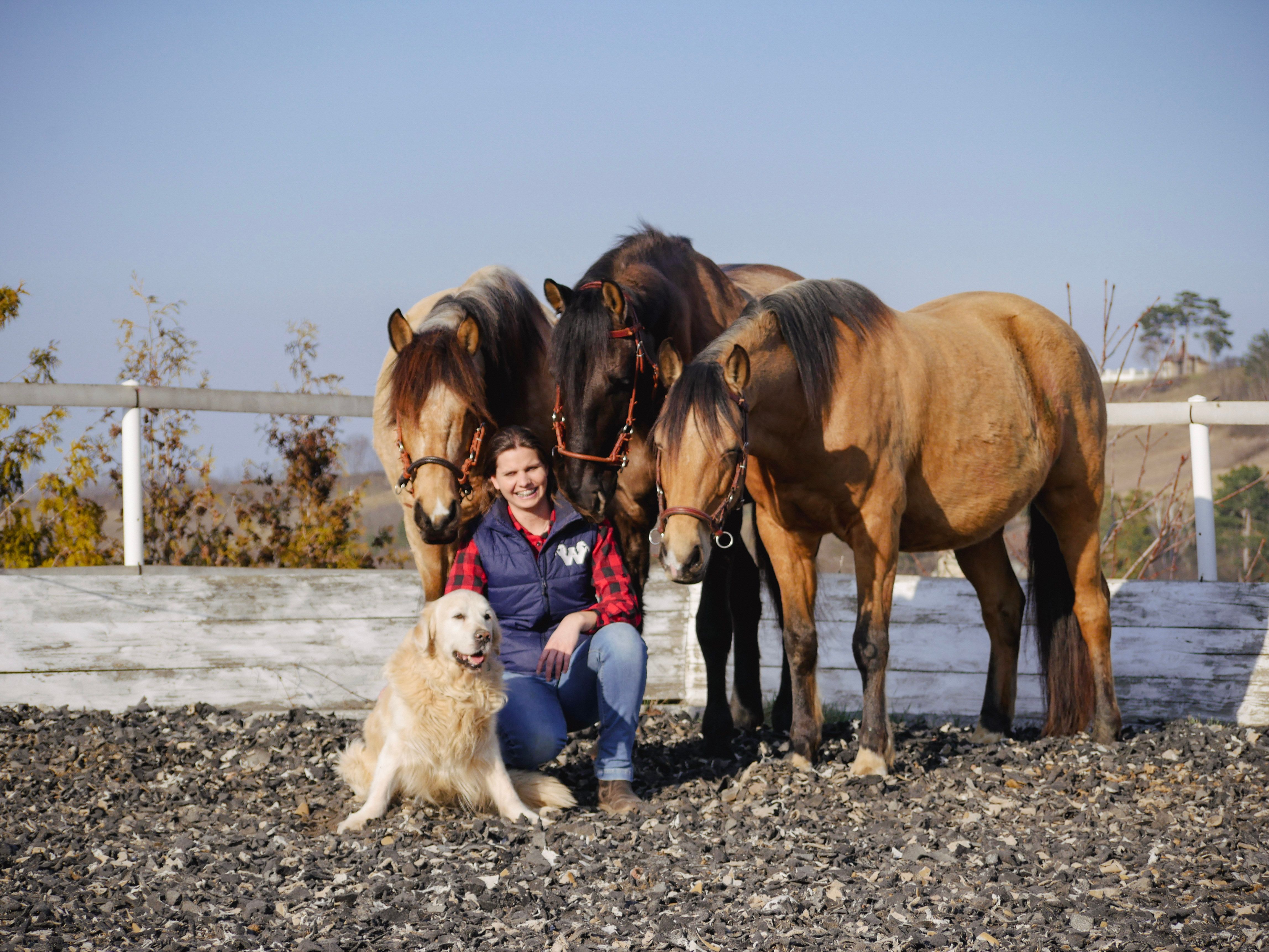 Eine Frau sitzt lächelnd auf einem Reitplatz, umgeben von drei Pferden und einem Hund.