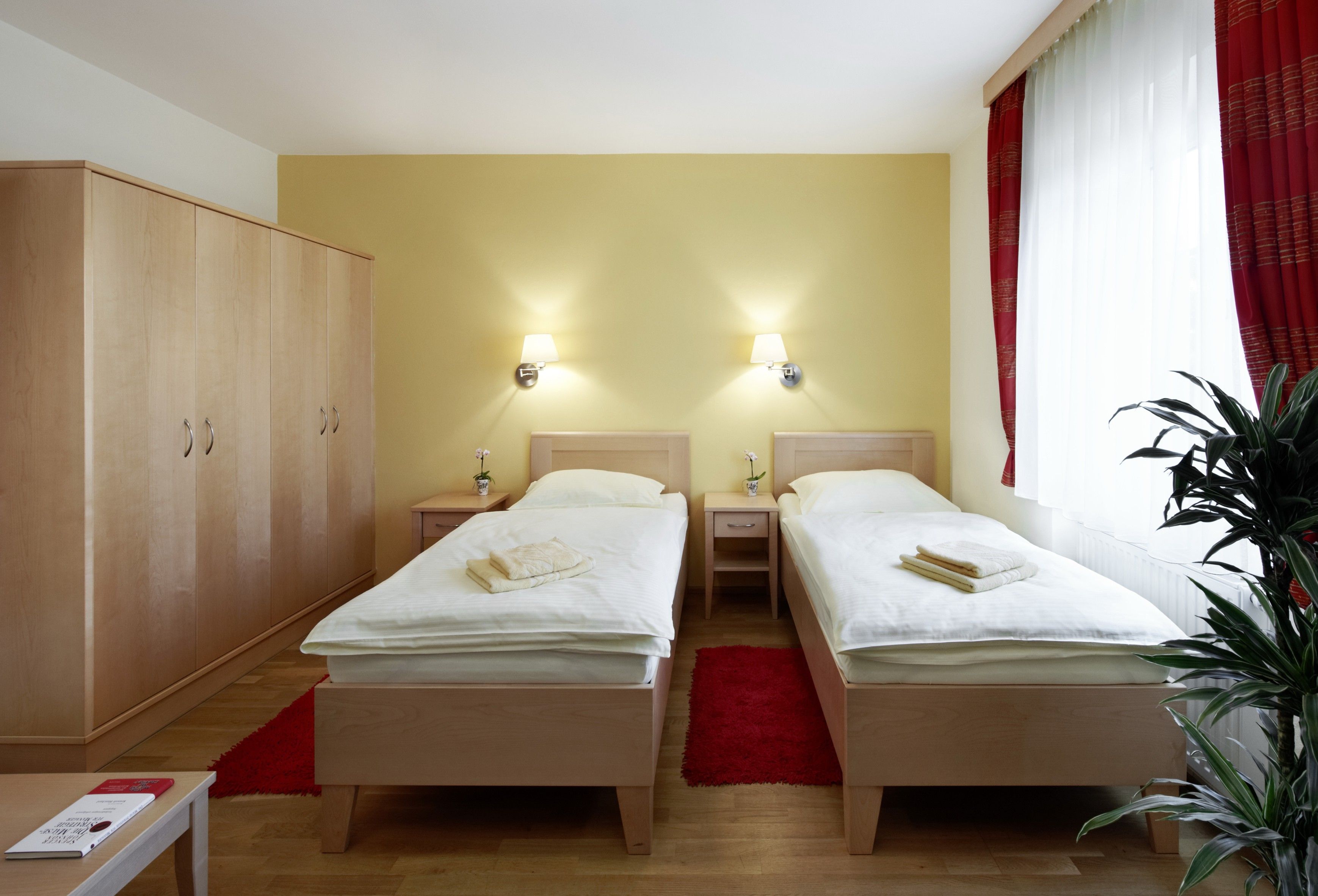 Ein Gästezimmer mit zwei Einzelbetten, einem großen Kleiderschrank, roten Vorhängen und einer Pflanze.