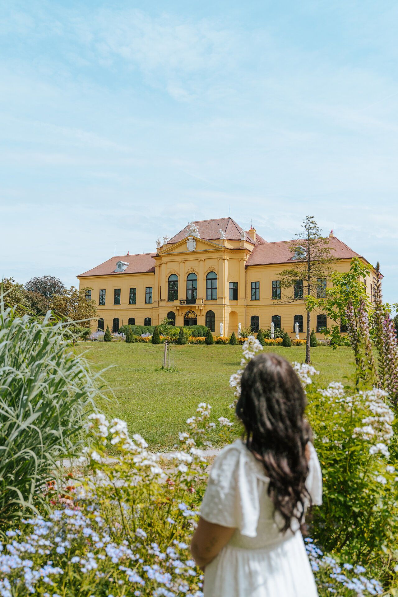 Umgeben von blühenden Blumen und üppigem Grün strahlt das Schloss eine majestätische Ruhe aus. Die sanften Hügel im Hintergrund laden zu einem entspannten Spaziergang durch den Schlosspark ein, während die warmen Sonnenstrahlen die Atmosphäre mit einem goldenen Glanz erfüllen.