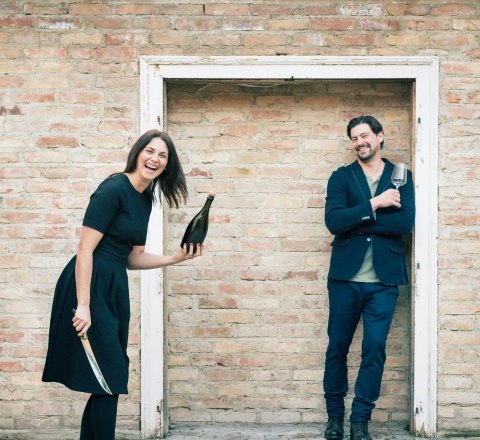 Zwei Personen vor einer Backsteinwand, eine hält eine Flasche und ein Messer, die andere ein Glas.