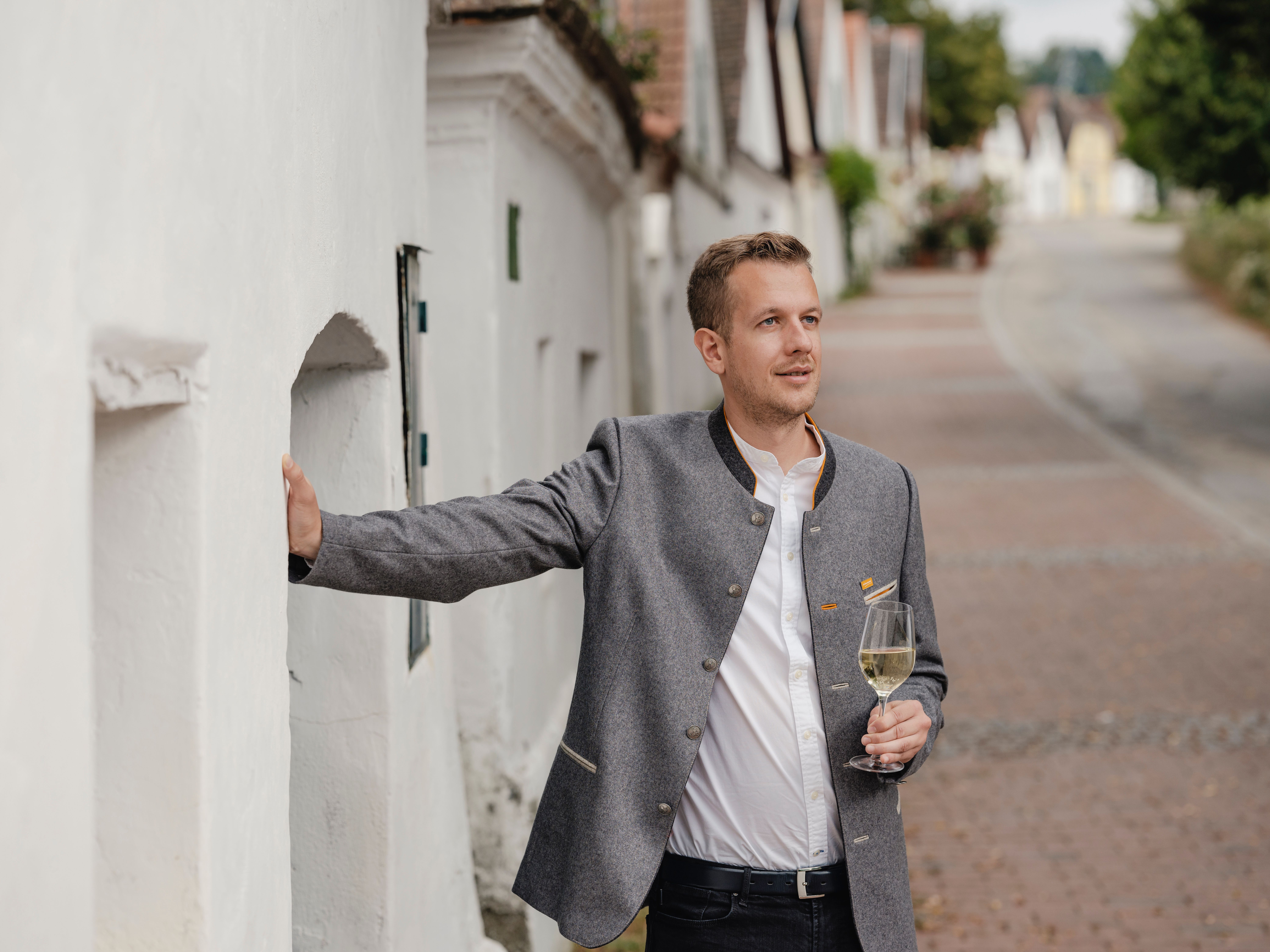 Ein Mann - in einen grauen Weinviertel-Trachtenjanker gekleidet - steht in einer Kellergasse, in der Hand hält er ein Glas Weißwein.