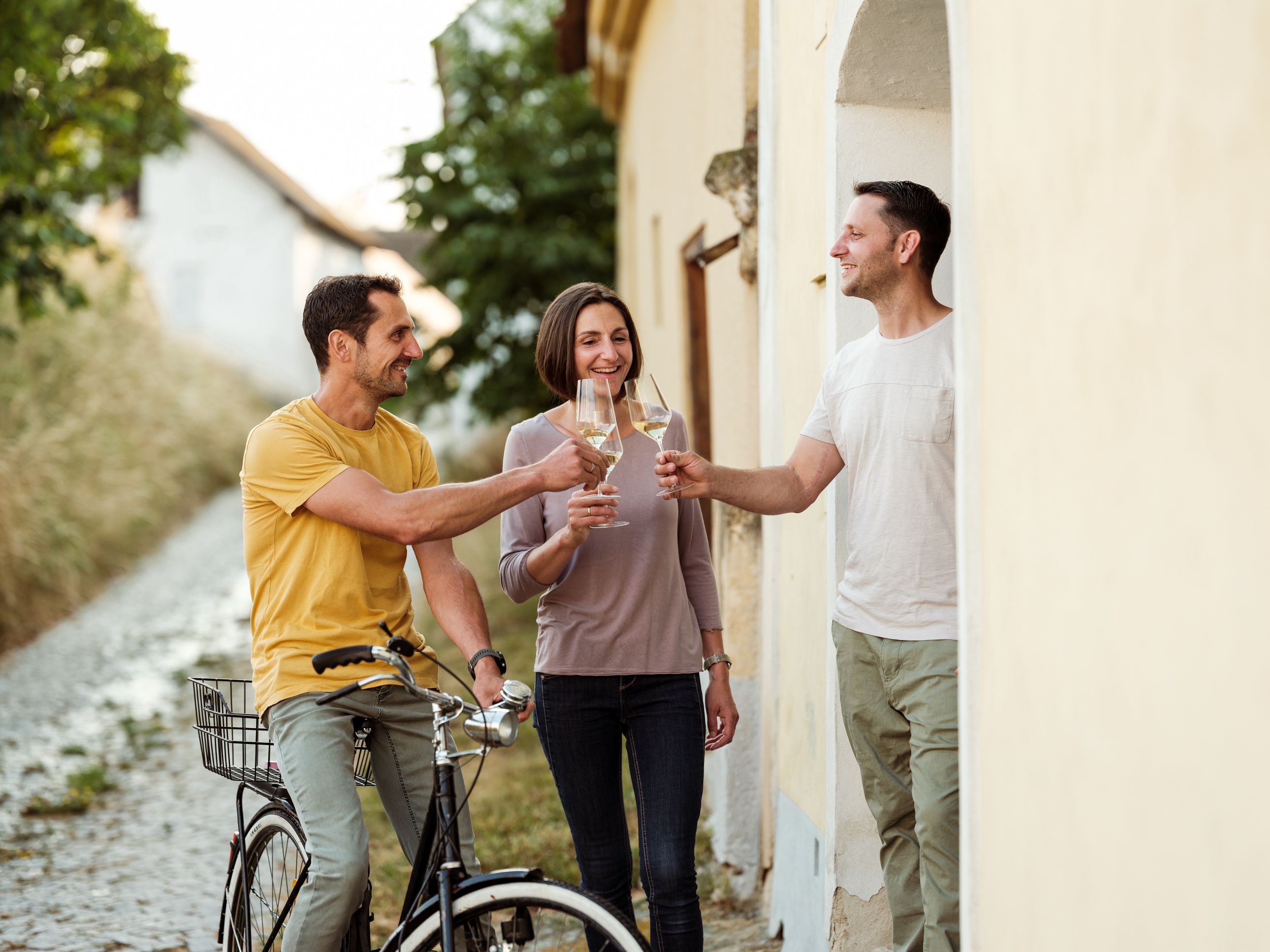 Drei Personen stoßen mit Weingläsern in einer ländlichen Gasse an.