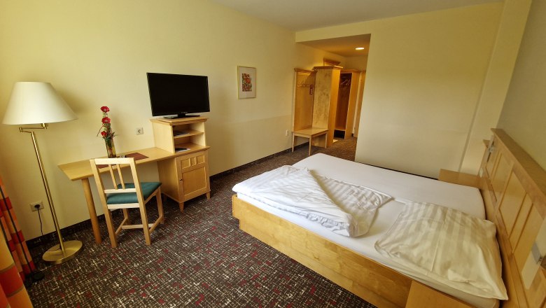 Ein einfach eingerichtetes Gästezimmer mit Bett, Schreibtisch, Stuhl, Fernseher und Stehlampe.