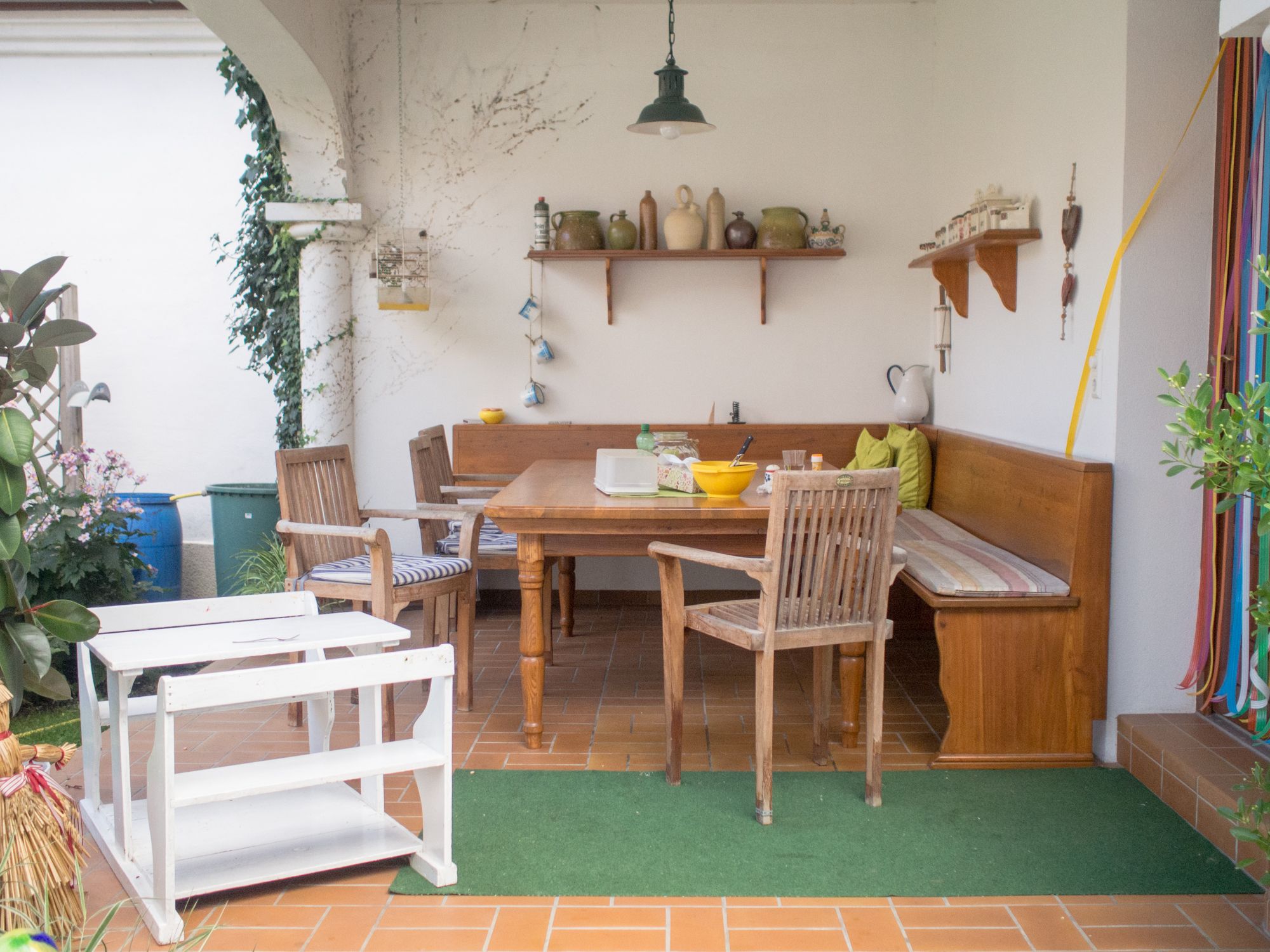Gemütliche Terrasse mit Holzmöbeln, Eckbank, Tisch und Dekorationen. Pflanzen und Regale mit Keramik im Hintergrund.