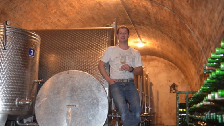 A man stands in a wine cellar next to large stainless steel tanks and bottles of wine.