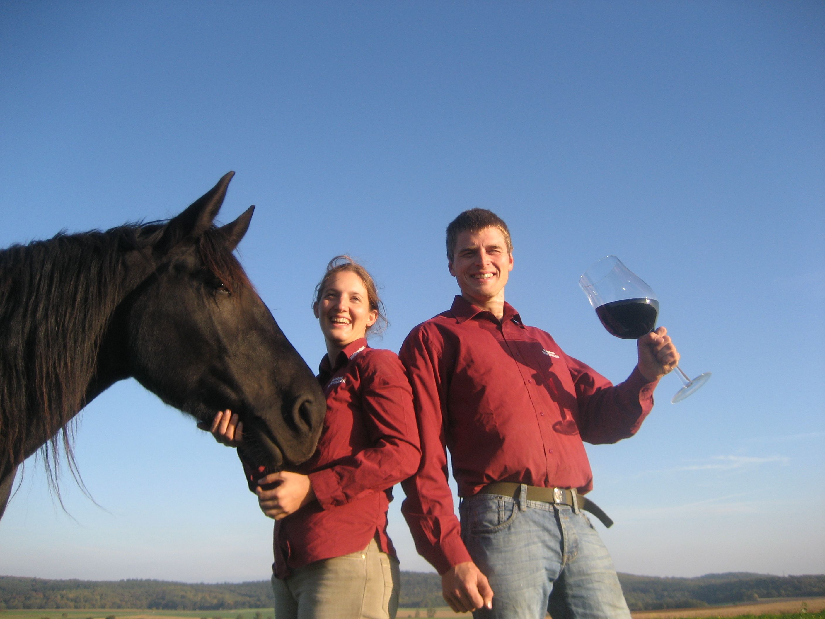 Zwei Personen in roten Hemden mit einem schwarzen Pferd und einem großen Weinglas im Freien.