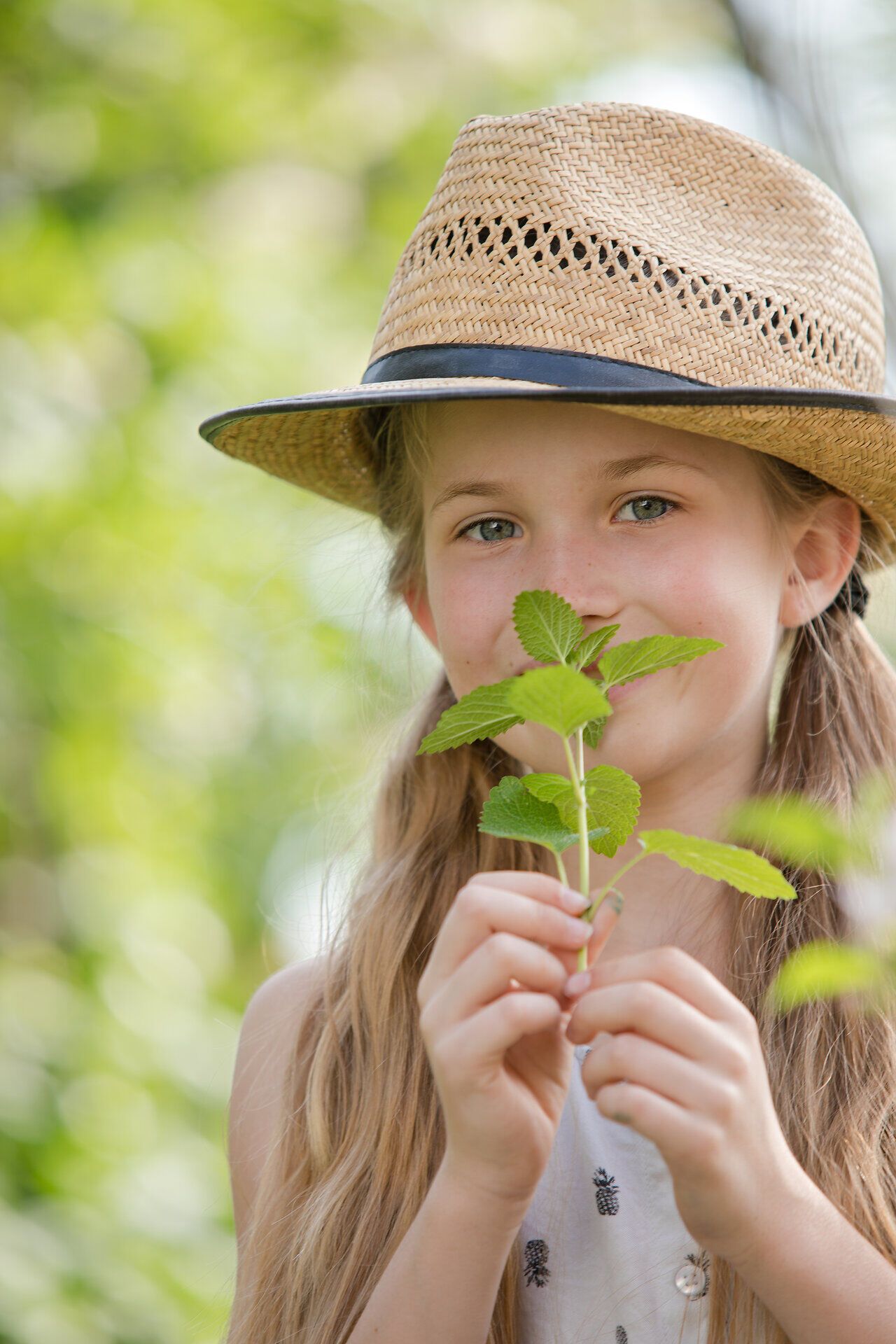 Ein fröhliches Kind mit einem Strohhut hält zarte Blätter in der Hand und genießt die warmen Sonnenstrahlen. Umgeben von üppigem Grün strahlt die Szene eine unbeschwerte Sommerfreude aus, die zum Entdecken der Natur einlädt.