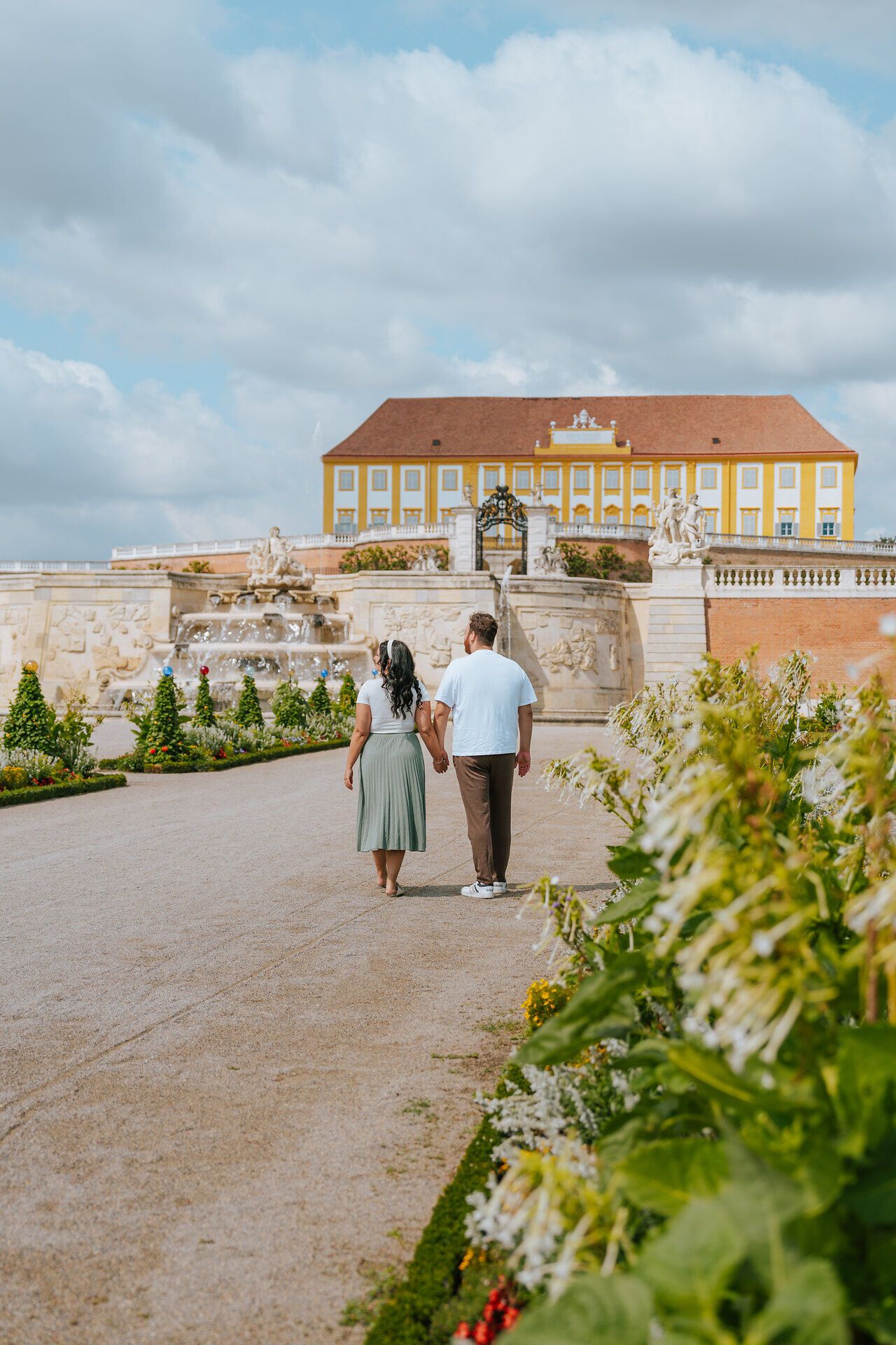 Ein romantisches Paar schlendert Hand in Hand durch die weitläufigen Terrassen des Schlosses Hof, umgeben von üppigen Blumenbeeten und dem sanften Plätschern der Wasserfontänen. Die strahlende Sonne und die malerische Kulisse schaffen eine unvergessliche Atmosphäre, die zum Verweilen einlädt.