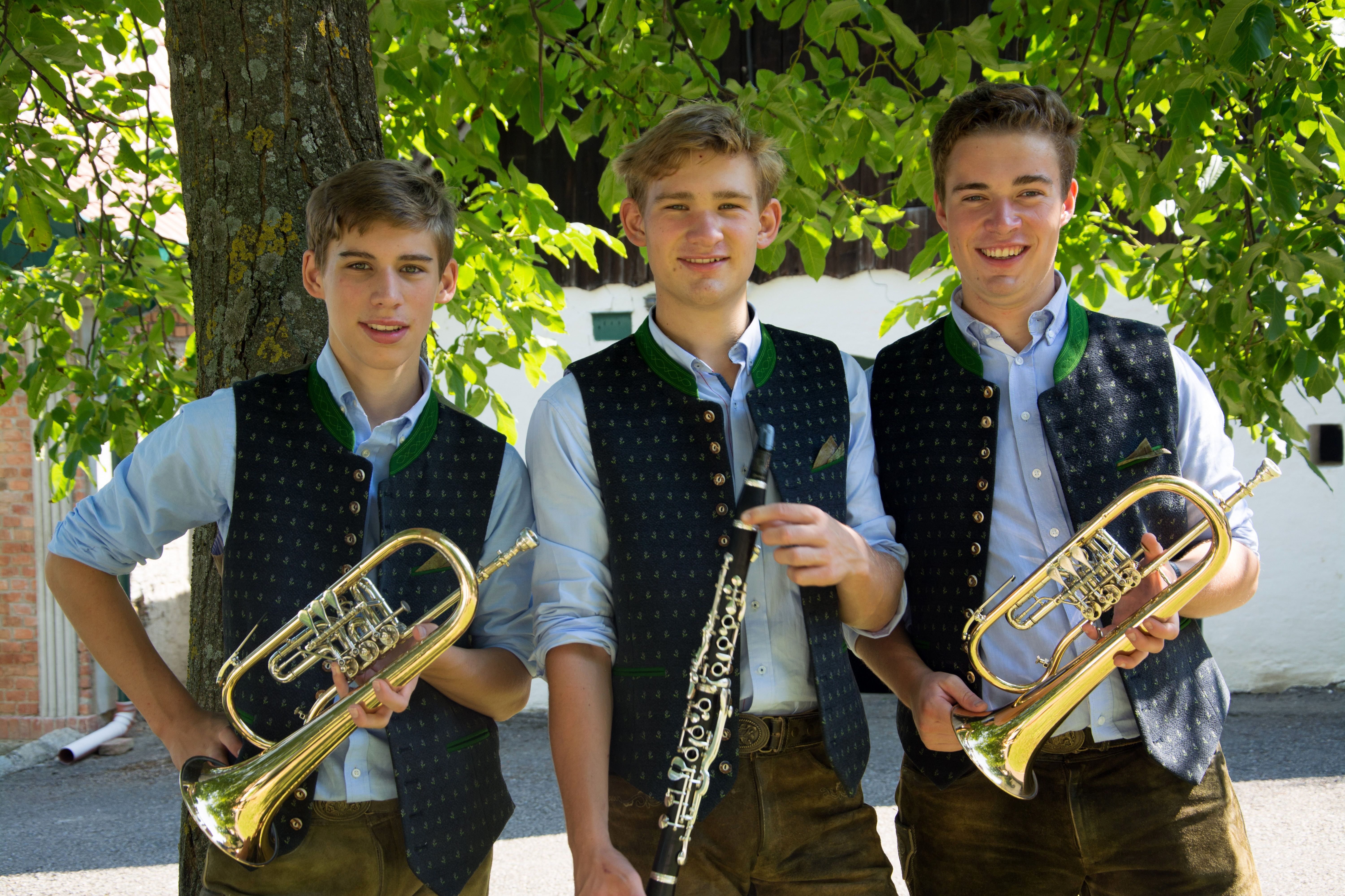 Three young men in traditional dress with wind instruments in front of a tree.