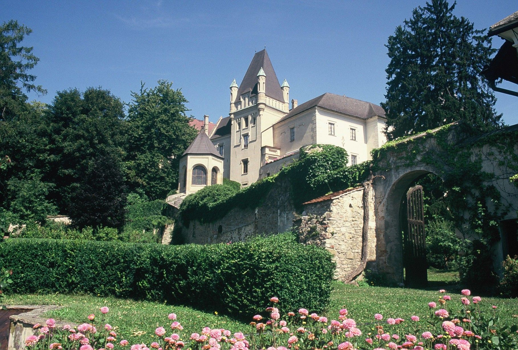 A castle with towers and a stone wall, surrounded by green trees and blossoming flowers in the foreground.