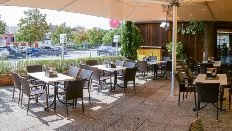 Ein sonniger Gastgarten mit leeren Tischen und Stühlen, umgeben von Pflanzen und einem großen Sonnenschirm.