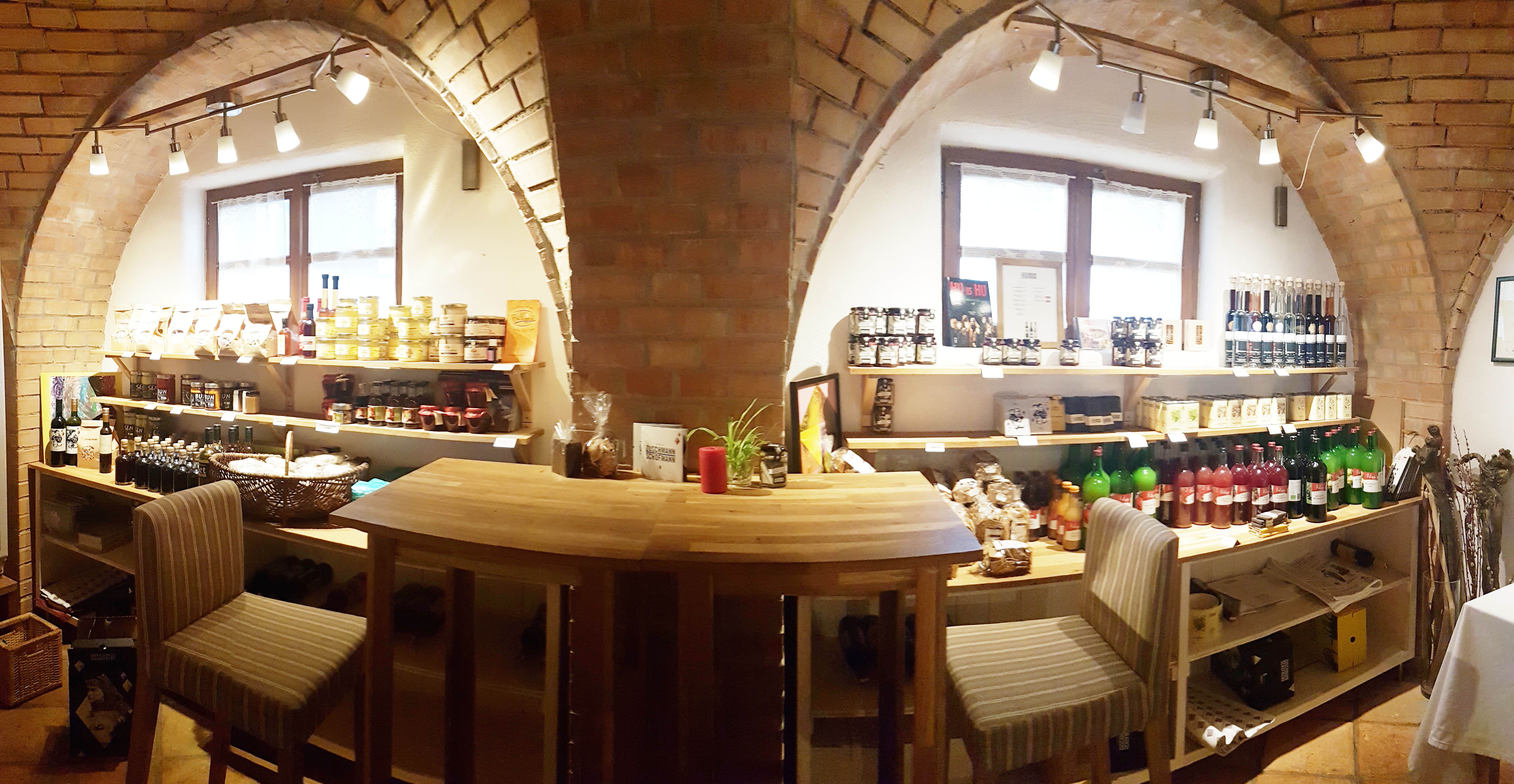Innenansicht eines kleinen Ladens mit Regalen voller Lebensmittel und Getränken, Holztisch und Stühlen.