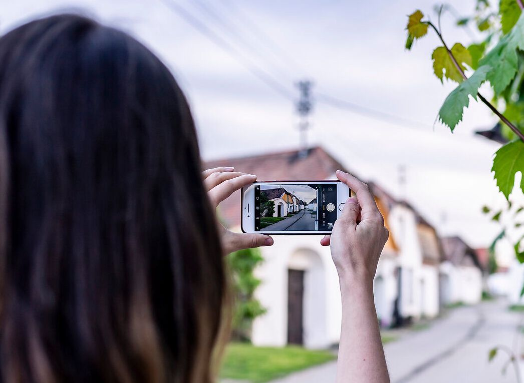 In der malerischen Kellergasse umgeben von üppigem Weinlaub, hält eine Person ihr Handy hoch, um die charmanten Weinhäuser festzuhalten. Die sanften Hügel im Hintergrund laden zu einem entspannten Spaziergang ein, während die frische Luft den Duft von reifen Trauben verbreitet.