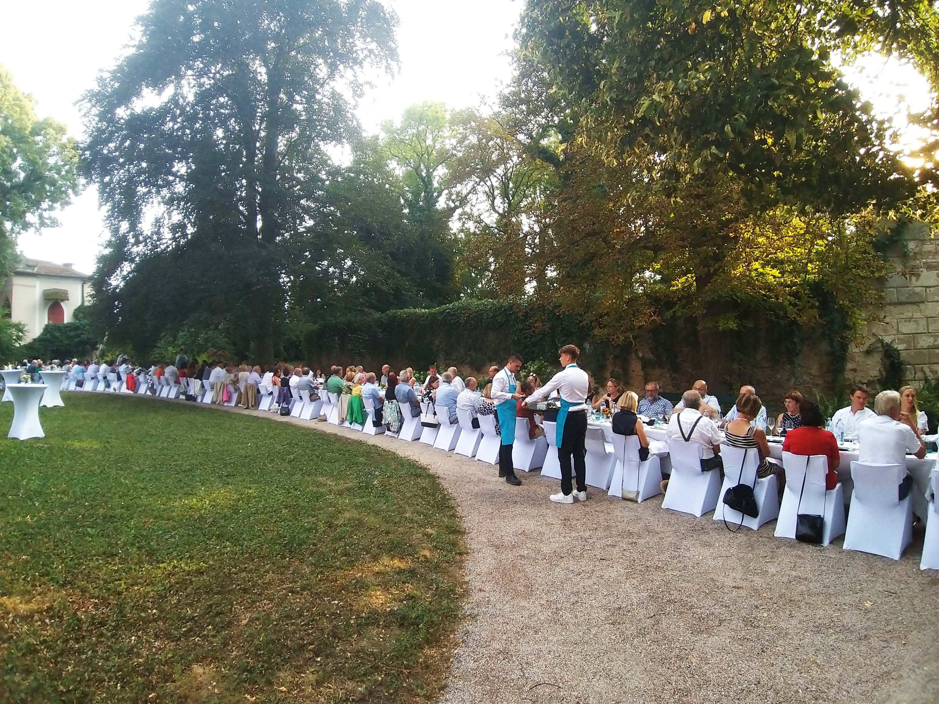 Langes Bankett im Freien mit vielen Gästen an weiß gedeckten Tischen, umgeben von Bäumen.