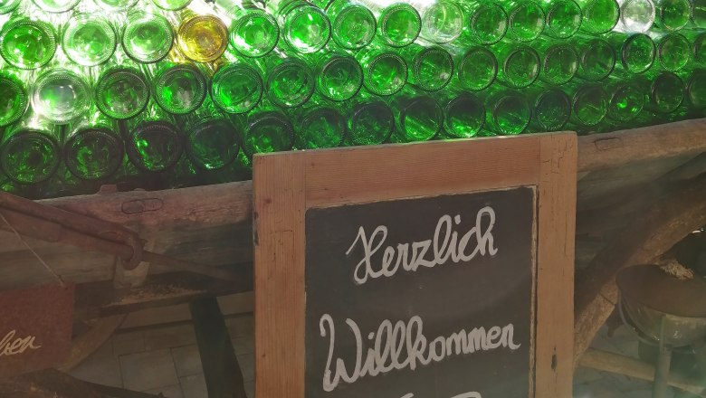 Ein Stapel grüner Glasflaschen auf einem Wagen mit einem Schild 'Herzlich Willkommen' davor, im Hintergrund ein Haus und Bäume.