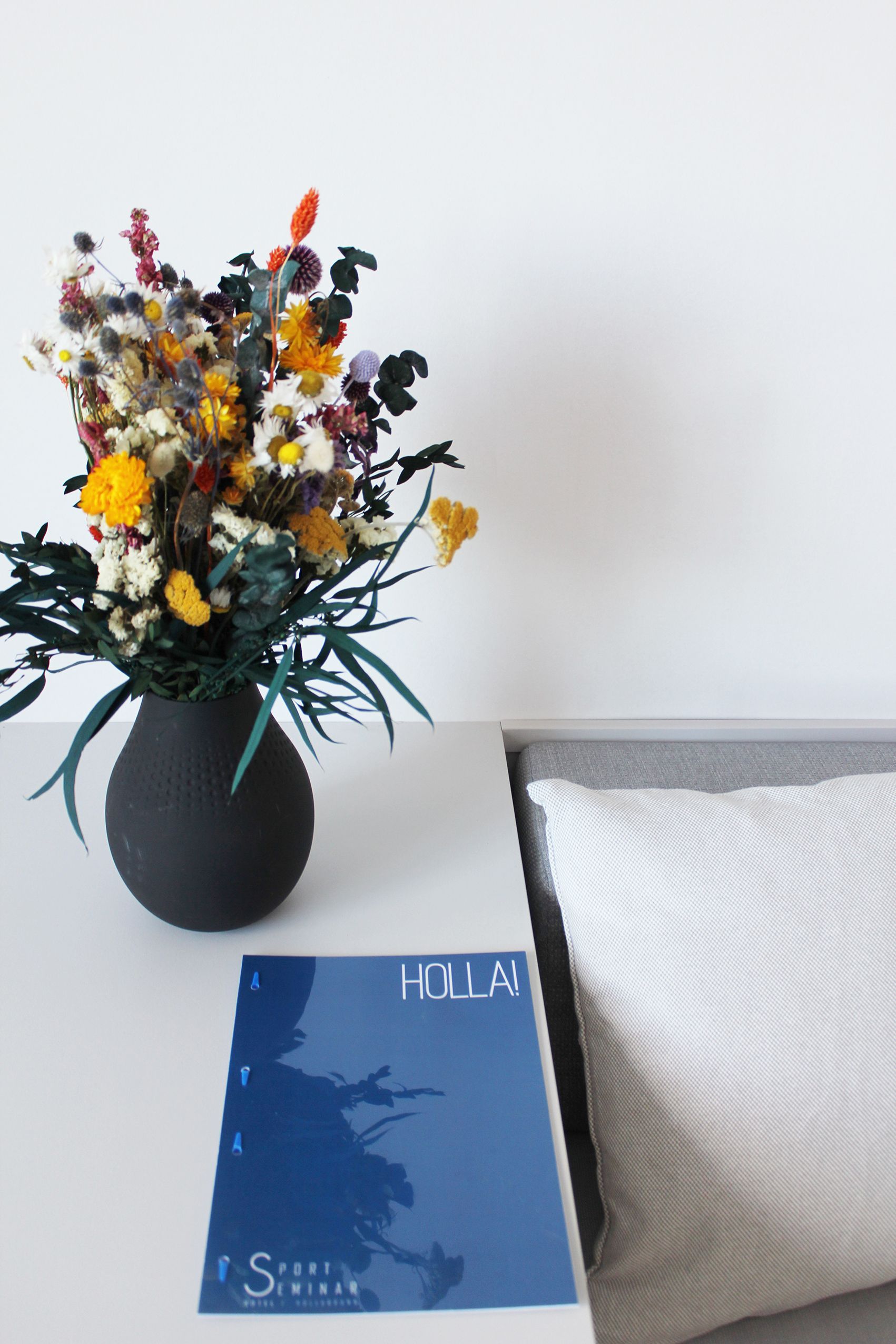 Ein Tisch mit einer schwarzen Vase voller bunter Blumen, einem blauen Magazin mit der Aufschrift 'HOLLA!' und einem weißen Kissen.
