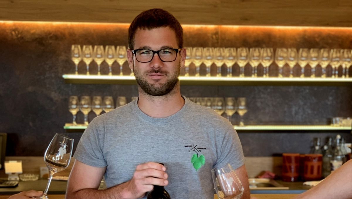 Ein Mann mit Brille hält eine Weinflasche und steht vor einem Regal mit Weingläsern.