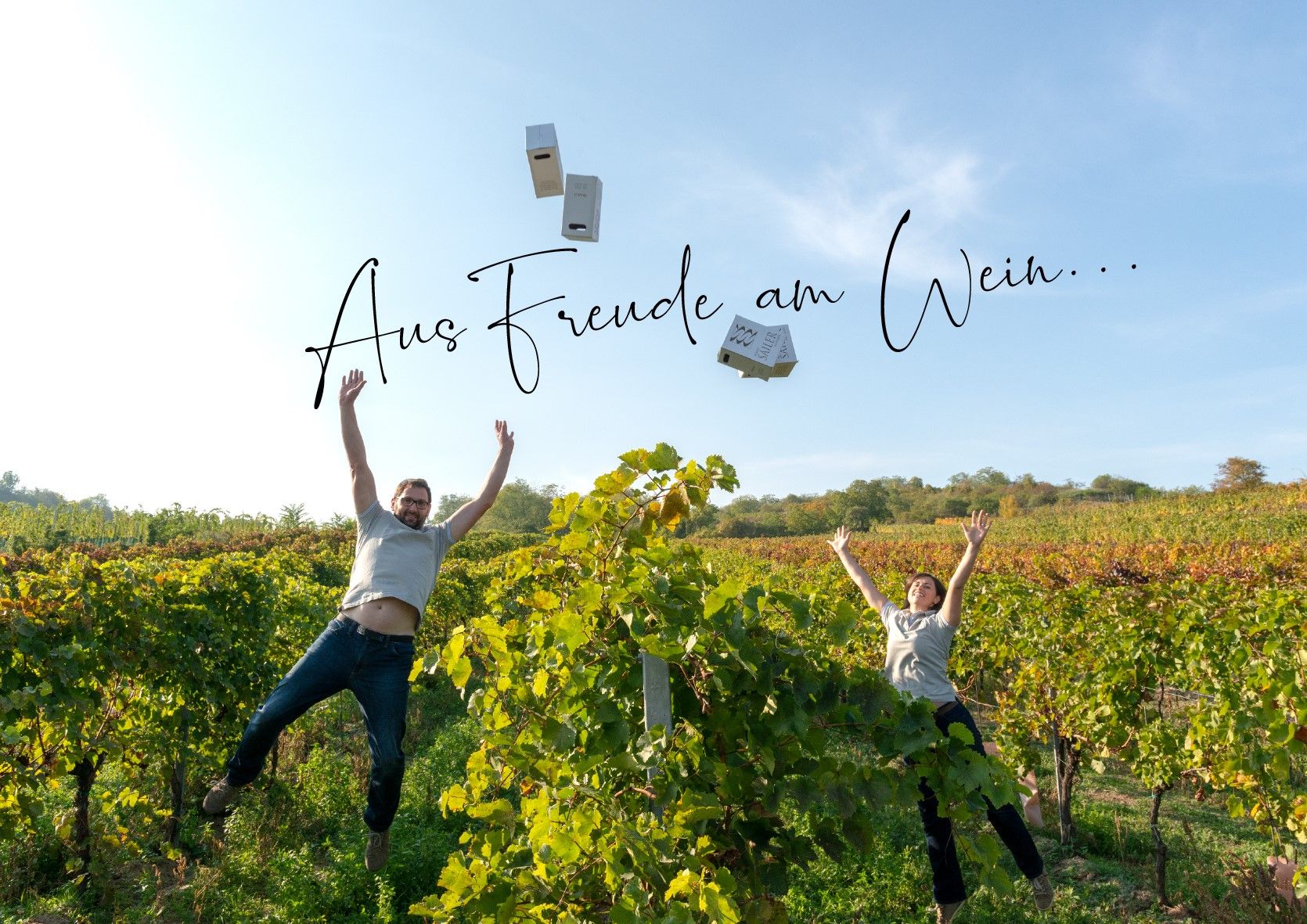 Zwei Personen springen in einem Weinberg in die Luft, während sie Kartons hochwerfen. Der Himmel ist blau und der Text 'Aus Freude am Wein...' ist zu sehen.