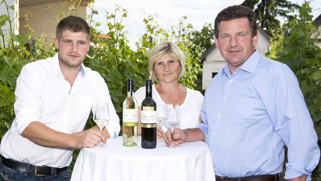 Drei Personen stehen an einem Tisch mit Weinflaschen im Freien.