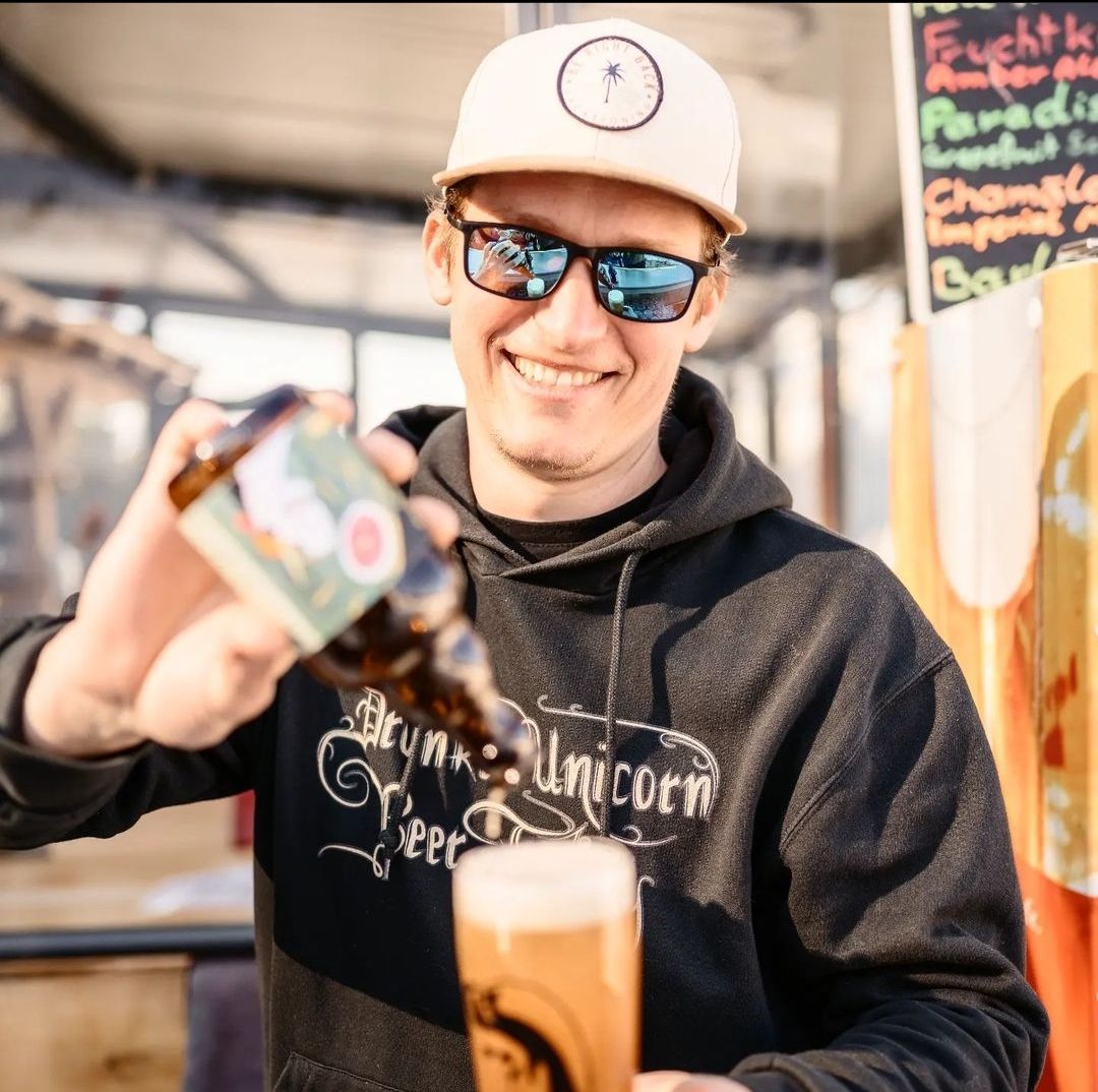 Man with sunglasses and cap pouring beer from a bottle into a glass.