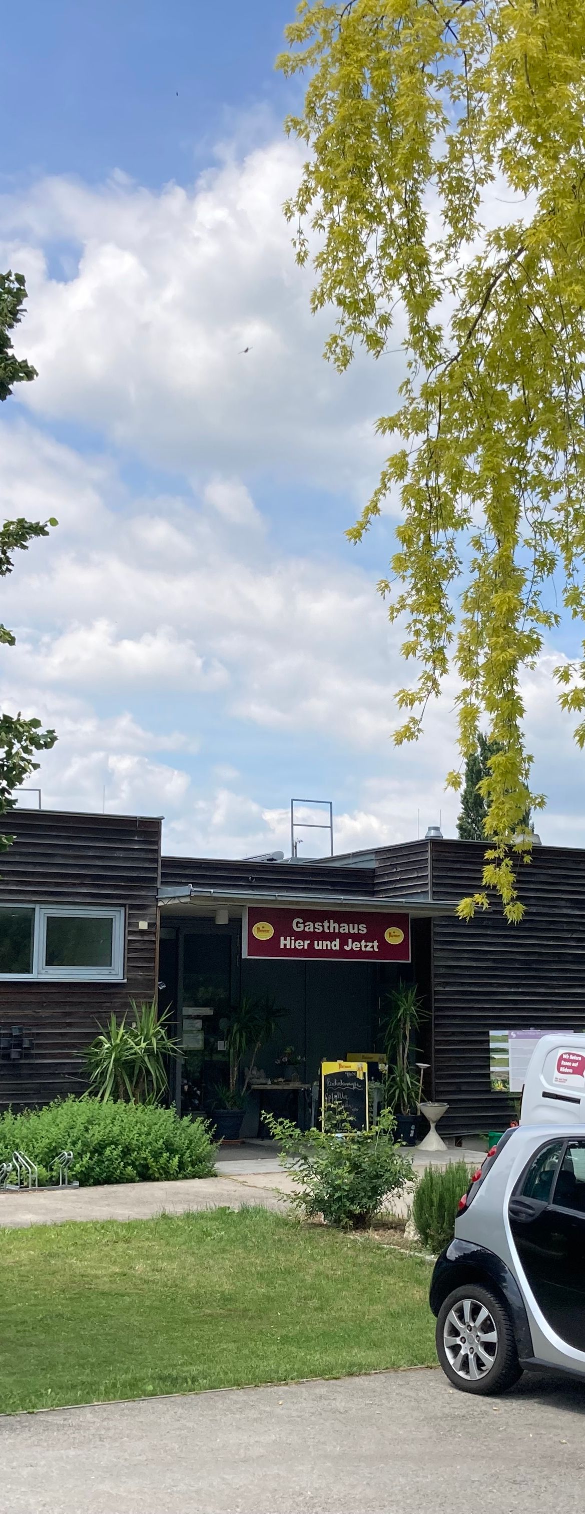 Blick durch Bäume auf ein Gebäude mit dem Schriftzug Gasthaus Hier und Jetzt und parkenden Autos im Vordergrund
