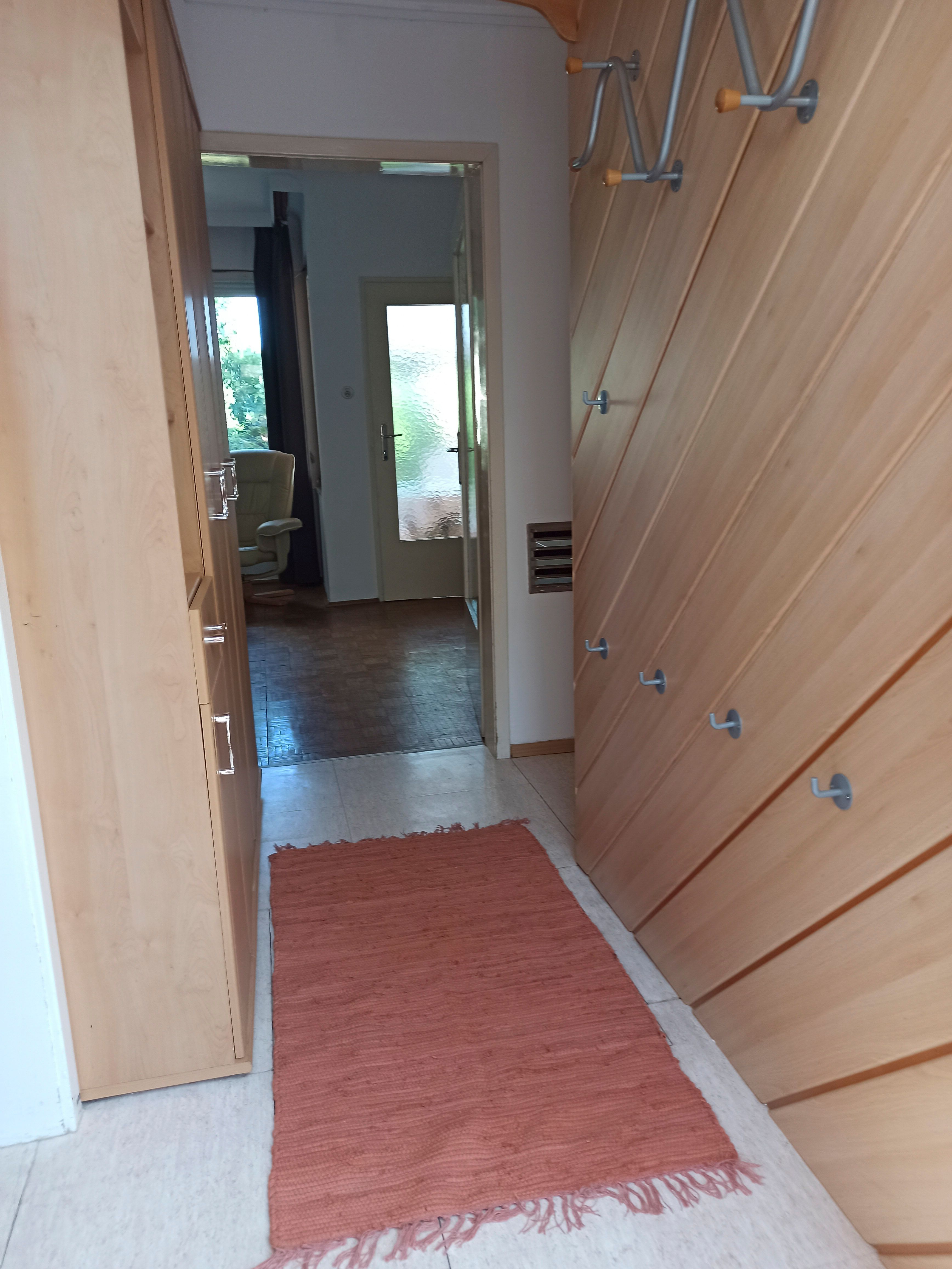 Ein Flur mit Holzwänden, Haken und einem roten Teppich. Im Hintergrund ist ein Raum mit einem Sessel und einer Glastür sichtbar.