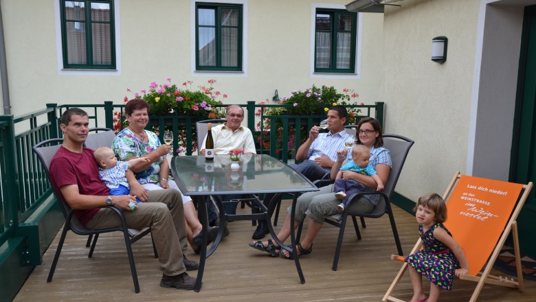 Eine Familie sitzt auf einer Terrasse an einem Tisch mit Getr&auml;nken. Im Hintergrund sind Blumen und ein Geb&auml;ude zu sehen.
