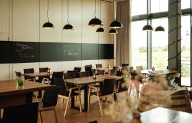Modernes Restaurant mit Holztischen, schwarzen St&uuml;hlen und gro&szlig;en Fenstern. Schwarze H&auml;ngelampen und eine Tafel an der Wand.