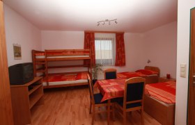 Ein einfach eingerichtetes Zimmer mit Etagenbett, Einzelbett, Tisch und Stühlen. Ein Fernseher steht auf einem Regal. Fenster mit Vorhängen im Hintergrund.