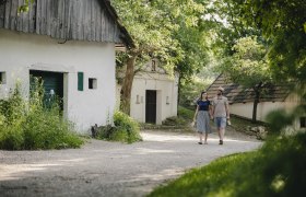 Ein Paar spaziert auf einem Weg zwischen alten, weißen Gebäuden in einer grünen, ländlichen Umgebung.