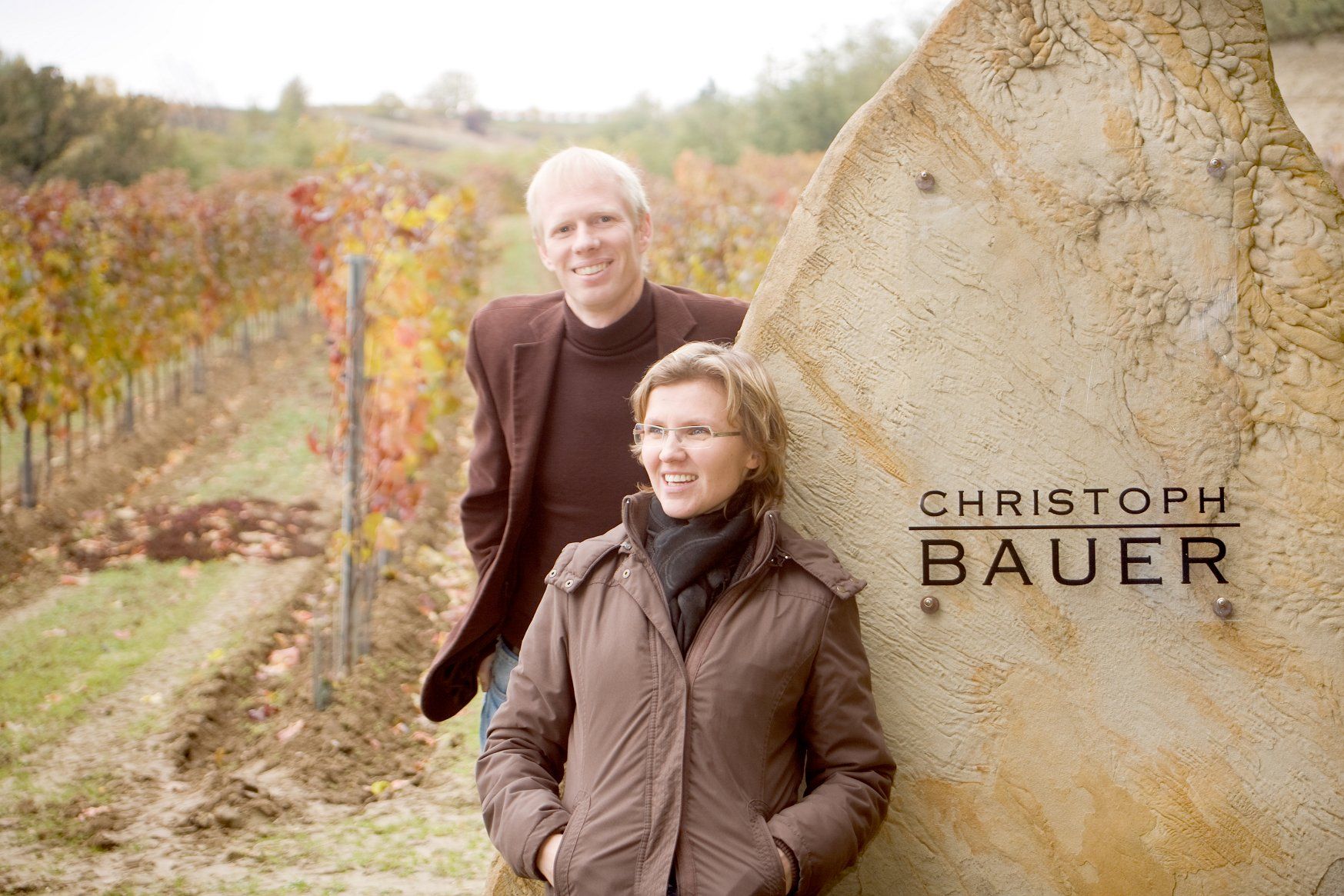 Zwei Personen stehen lächelnd vor einem Weinberg neben einem Stein mit der Aufschrift 'Christoph Bauer'.