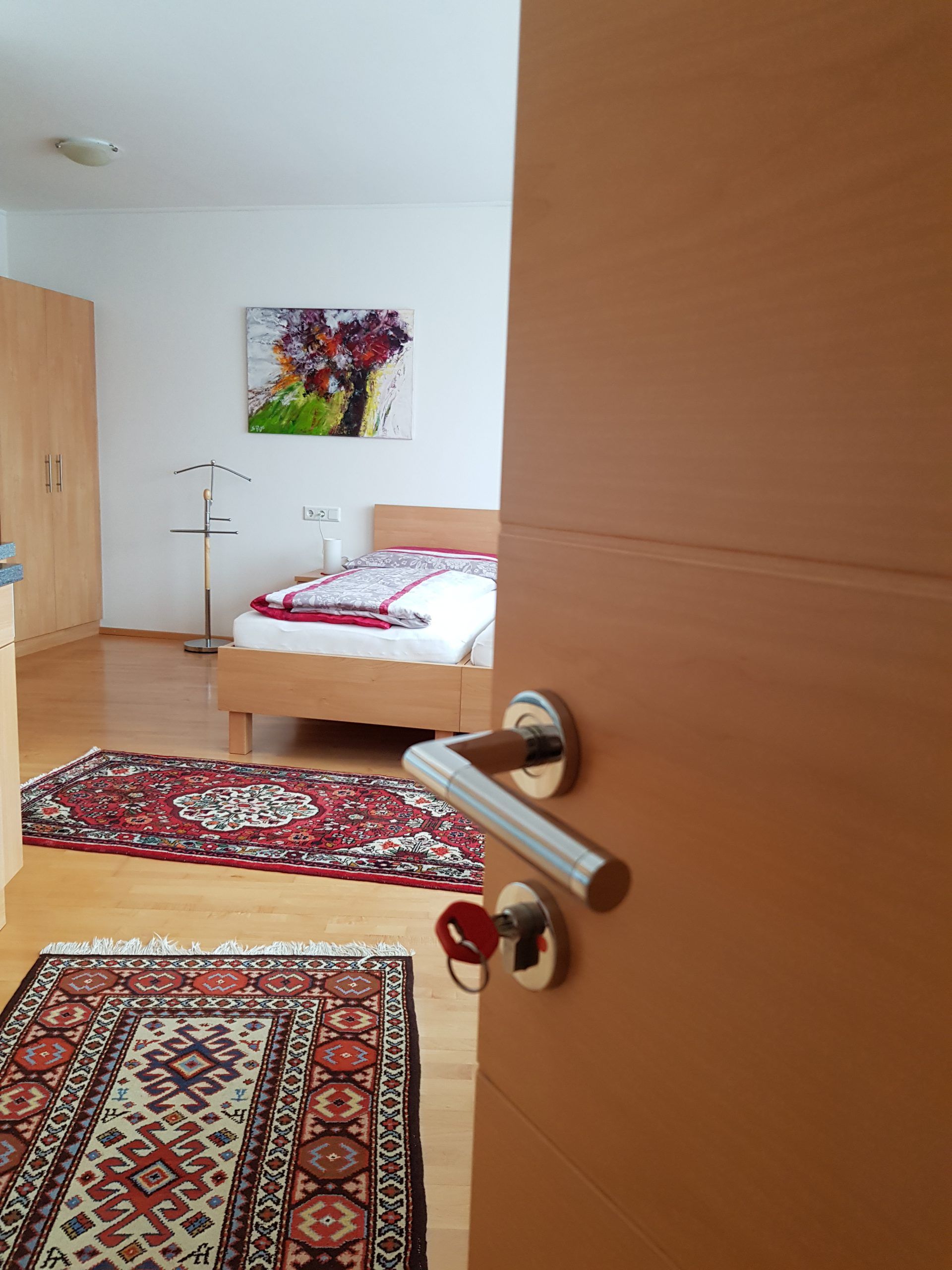 Ein geöffnetes Zimmer mit einem Bett, Teppichen und einem Gemälde an der Wand.