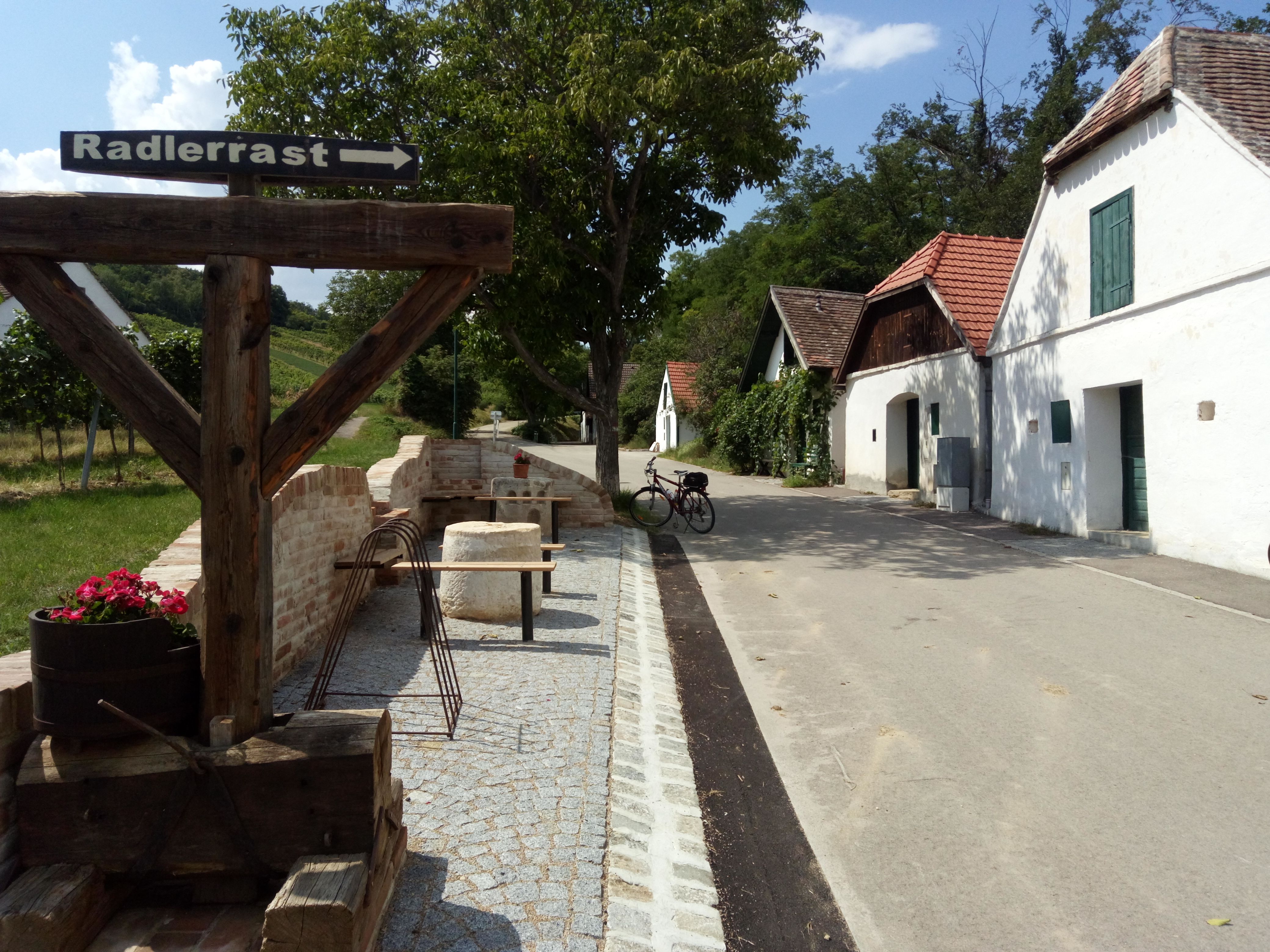 Ein Rastplatz für Radfahrer in Mailberg mit einem Holzschild, das auf die Radlerrast hinweist. Im Hintergrund sind Weinkeller und ein Fahrrad zu sehen.