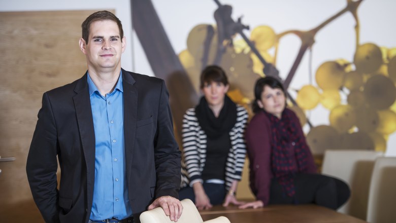 Ein Mann in einem blauen Hemd und schwarzem Sakko steht vor einem Tisch, im Hintergrund sitzen zwei Frauen.