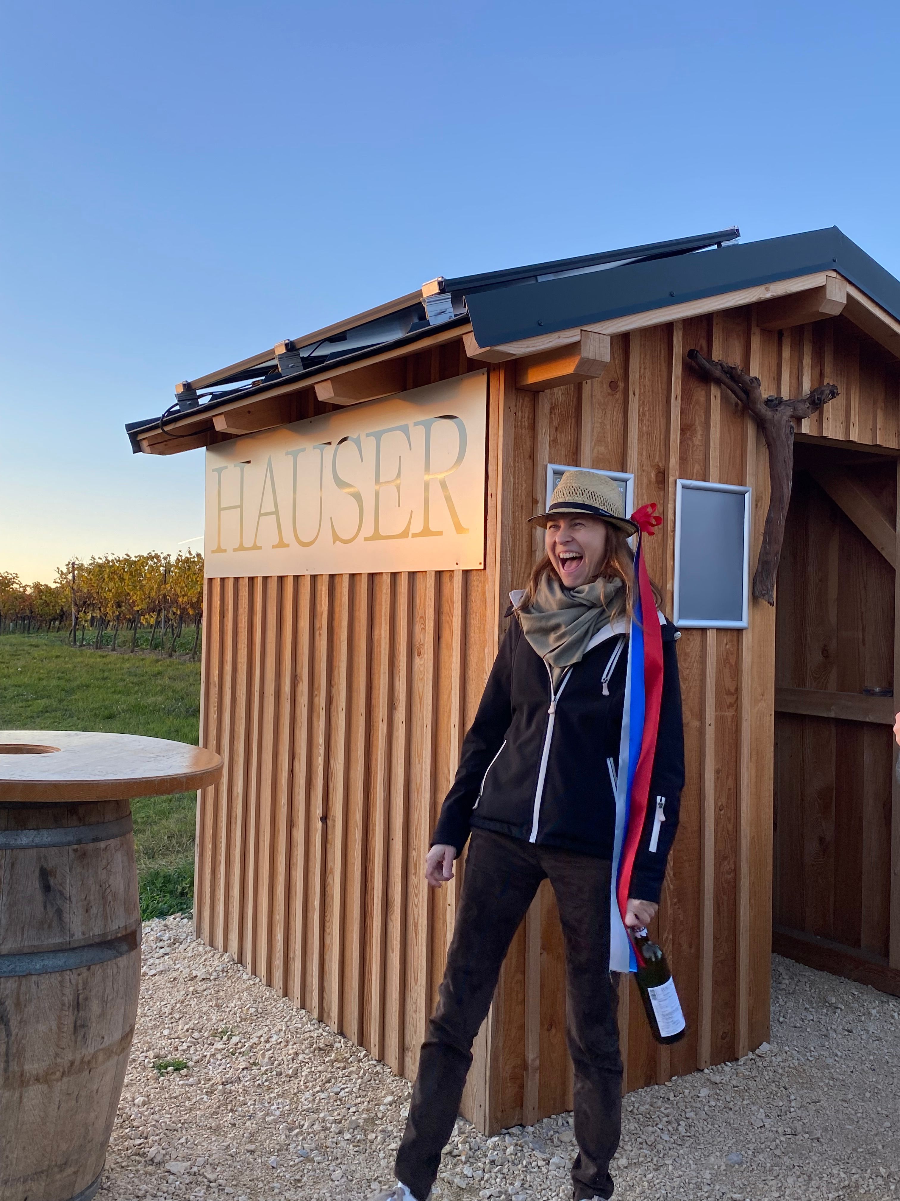Eine Person mit Hut und Flasche steht vor einer Holzhütte mit der Aufschrift 'Hauser'.