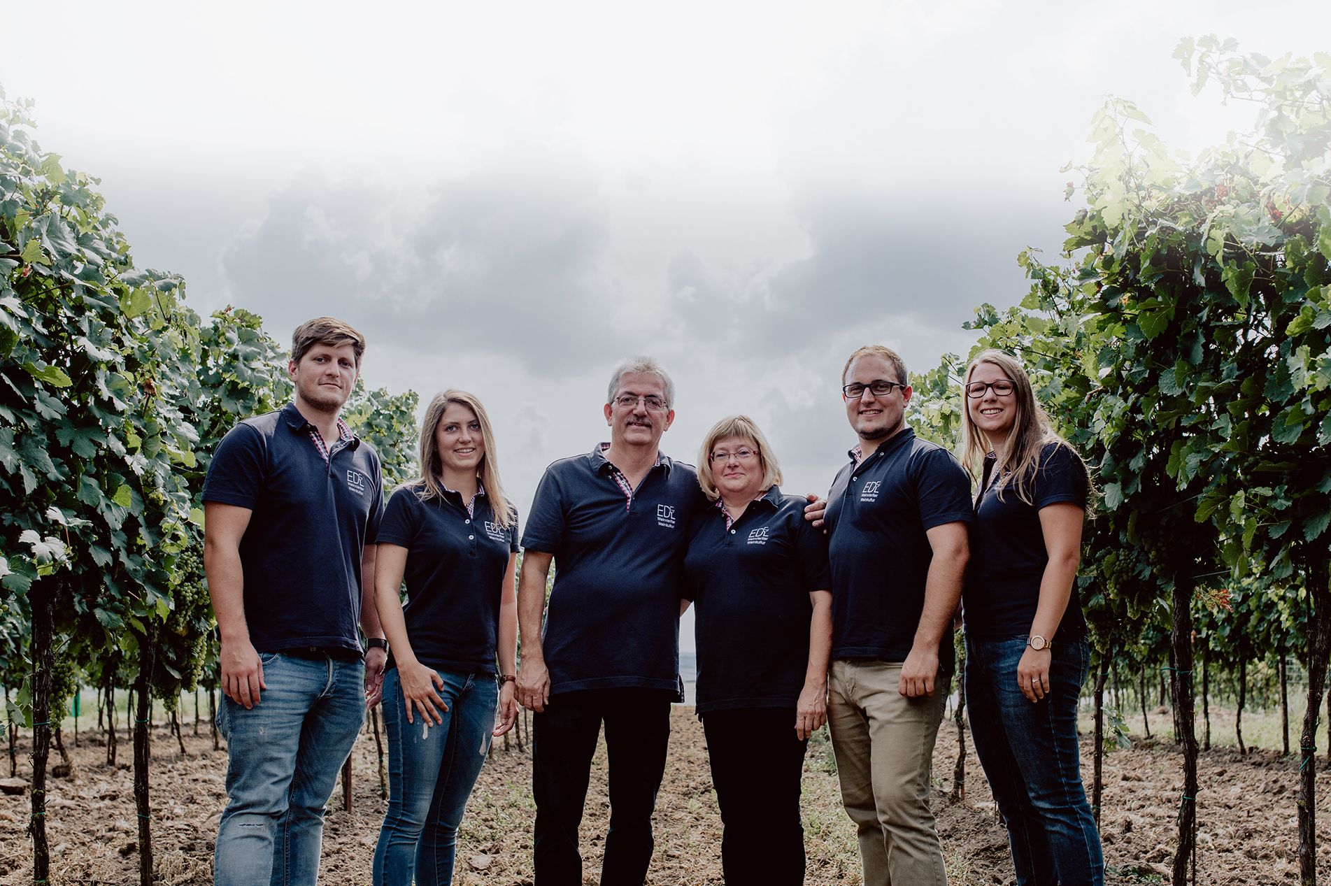 A family is standing in a vineyard, all wearing dark blue polo shirts with a logo.