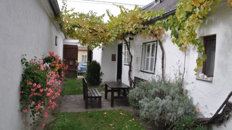 Ein gemütlicher Innenhof mit Holztisch und Bänken, umgeben von weißen Wänden, Blumen und Weinreben.