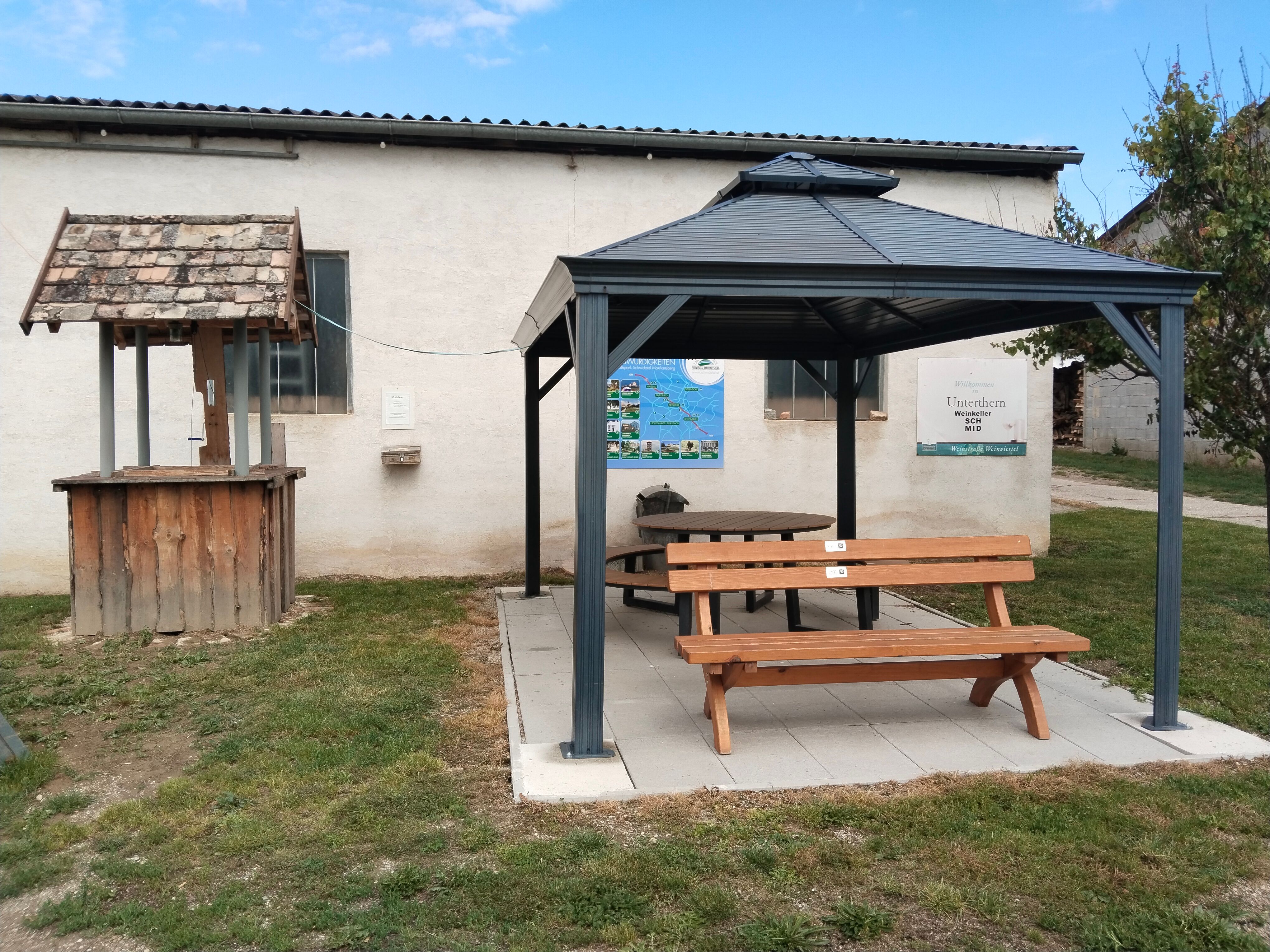 Ein Holzbrunnen und ein Pavillon mit Sitzbank vor einem Gebäude mit Schildern und Plakaten.