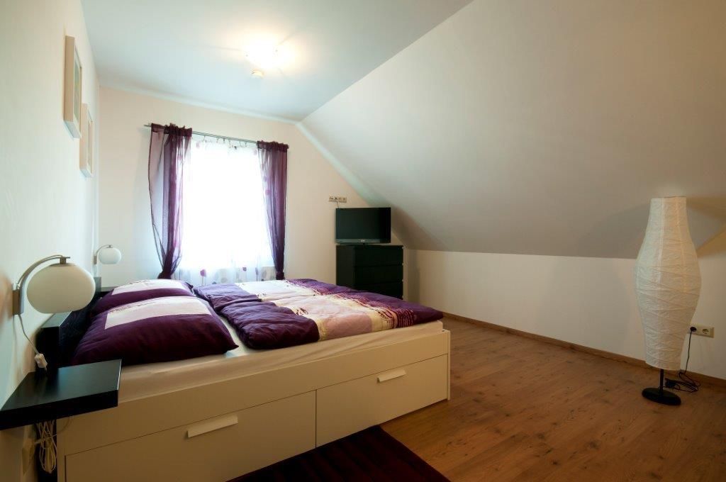 Ein gemütliches Gästezimmer mit einem Doppelbett, lila Vorhängen und einem Fernseher auf einer Kommode unter einer Dachschräge.