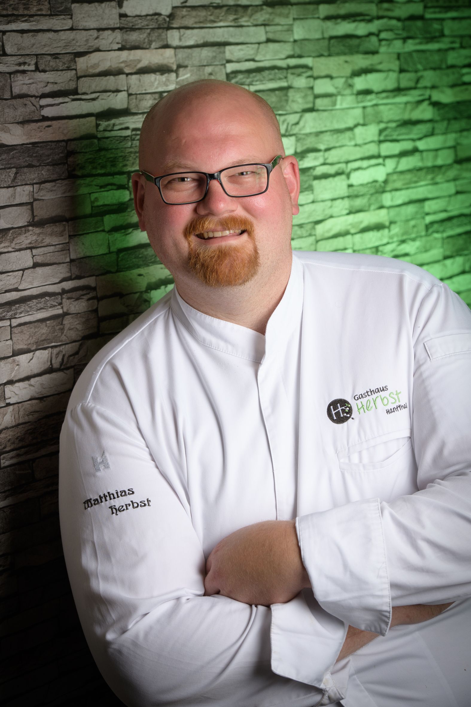 Ein Mann in weißer Kochjacke mit dem Logo 'Gasthaus Herbst' steht vor einer Steinwand.