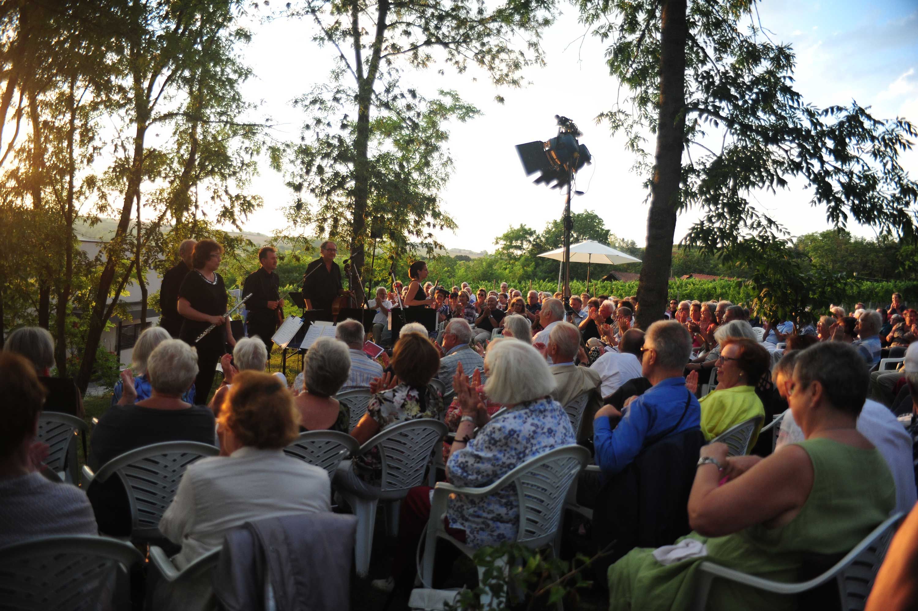 An outdoor concert with an orchestra and audience at sunset.