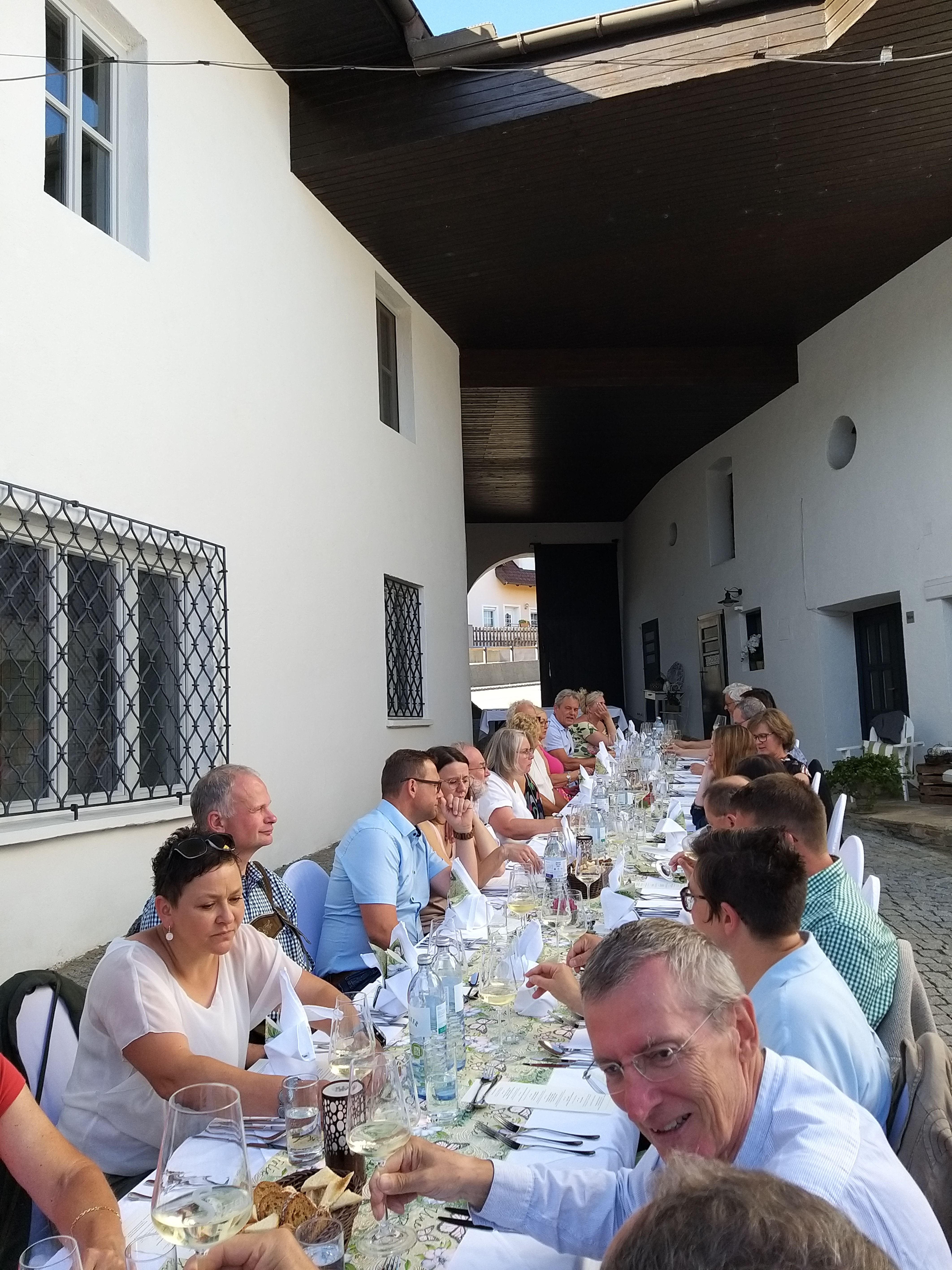 Menschen sitzen an einem langen Tisch im Freien und genießen ein gemeinsames Essen.