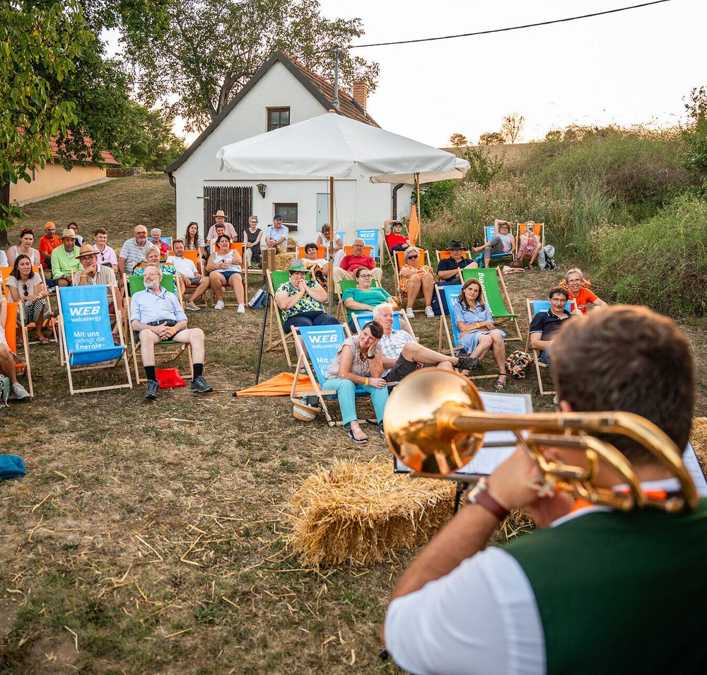 Unter dem sanften Licht der Abendsonne versammeln sich fröhliche Menschen auf dem Sierndorfer Kellerberg, um die Klänge des Wienerlied Festivals zu genießen. Die entspannte Atmosphäre wird von bunten Stühlen und der malerischen Kulisse der Weinregion umrahmt, während die Gäste in geselliger Runde die Kultur und Traditionen des Weinviertels feiern.