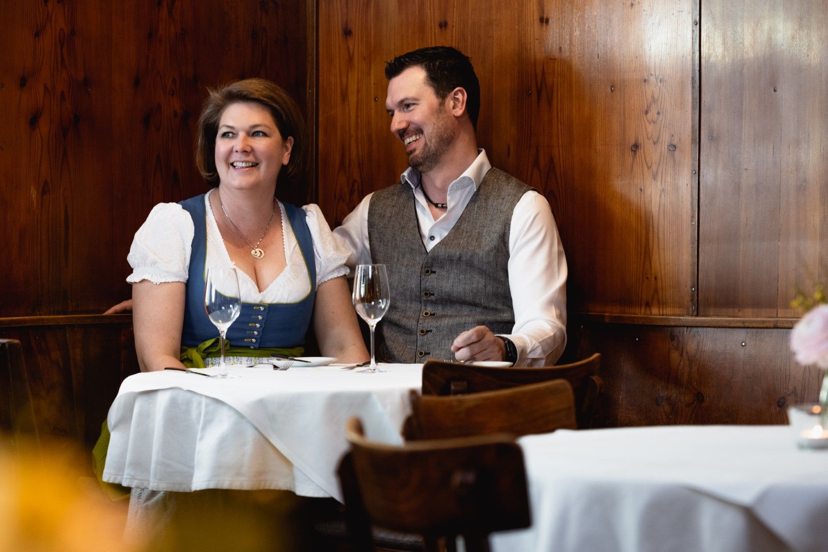 Ein Paar in traditioneller Kleidung sitzt lächelnd an einem Tisch in einem holzgetäfelten Raum.