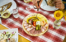 Ein gedeckter Tisch mit Aufschnittplatte, Brot, Wein und Blumen auf rot-weiß karierter Tischdecke.