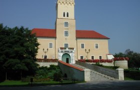 Ein Schloss mit einem Turm und roten D&auml;chern, umgeben von Blumen und B&auml;umen, unter einem klaren blauen Himmel.