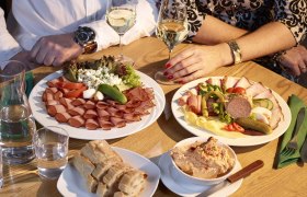Zwei Personen sitzen an einem Tisch mit Wurst- und Käseplatten, Brot und Wein.