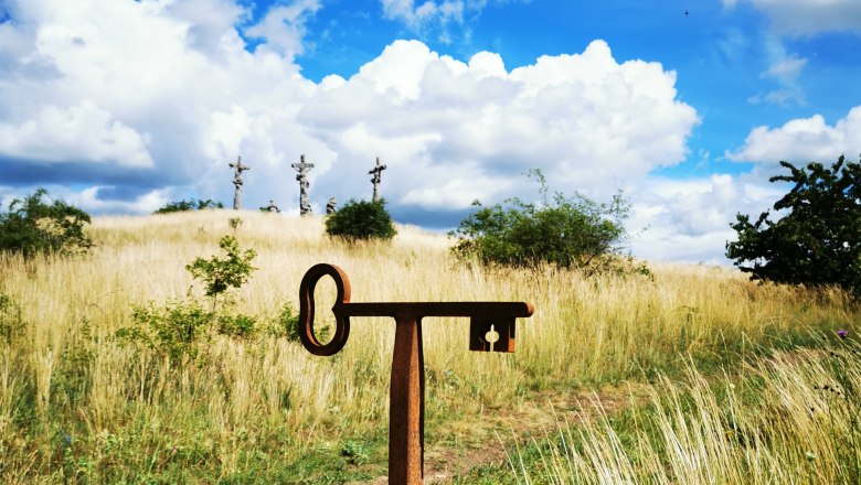 Ein Fotopunkt-Schild vor einer Wiese mit drei Kreuzen im Hintergrund unter blauem Himmel mit Wolken.