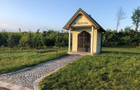 Kleine Kapelle mit Kreuz und Schriftzug "St. Hubertus" auf einer Wiese, mit gepflastertem Weg und Bank.